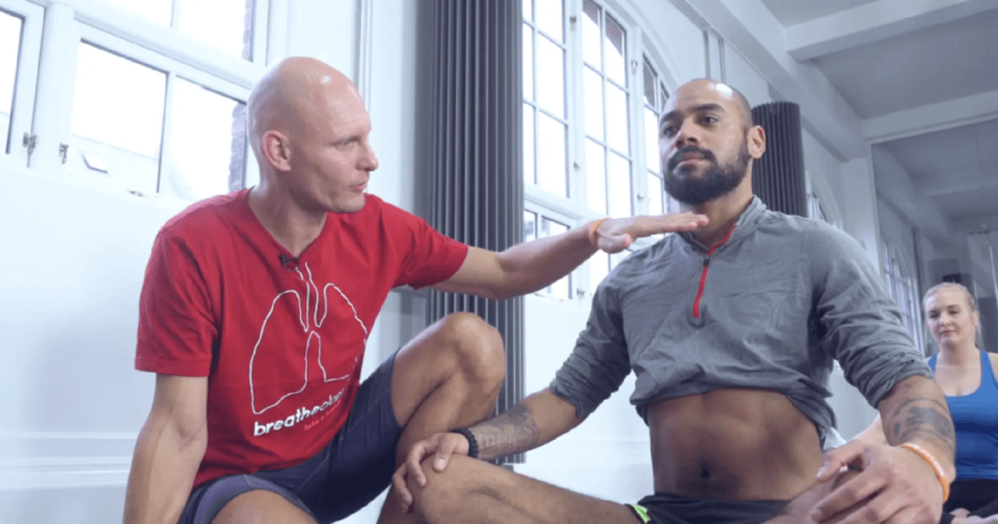 Stig Severinsen in a red Breatheology shirt guiding another man in a seated breathing exercise, with his hand placed near the person's chest in an indoor setting.