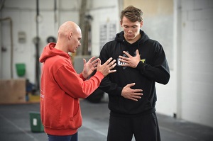 Stig Severinsen in a red hoodie instructing a man in a black hoodie on breathing techniques, with one hand on his chest and the other on his abdomen in a gym setting.