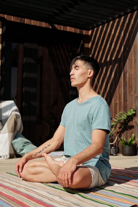 Man sitting cross-legged on a striped mat outdoors, meditating with eyes closed, and sunlight casting shadows in the background.