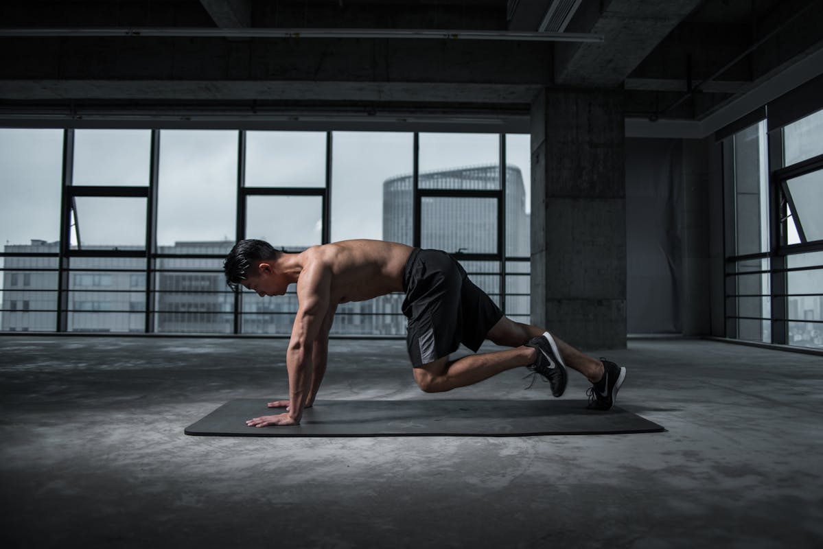 Shirtless man performing a mountain climber exercise on a yoga mat in an industrial-style gym with large windows.