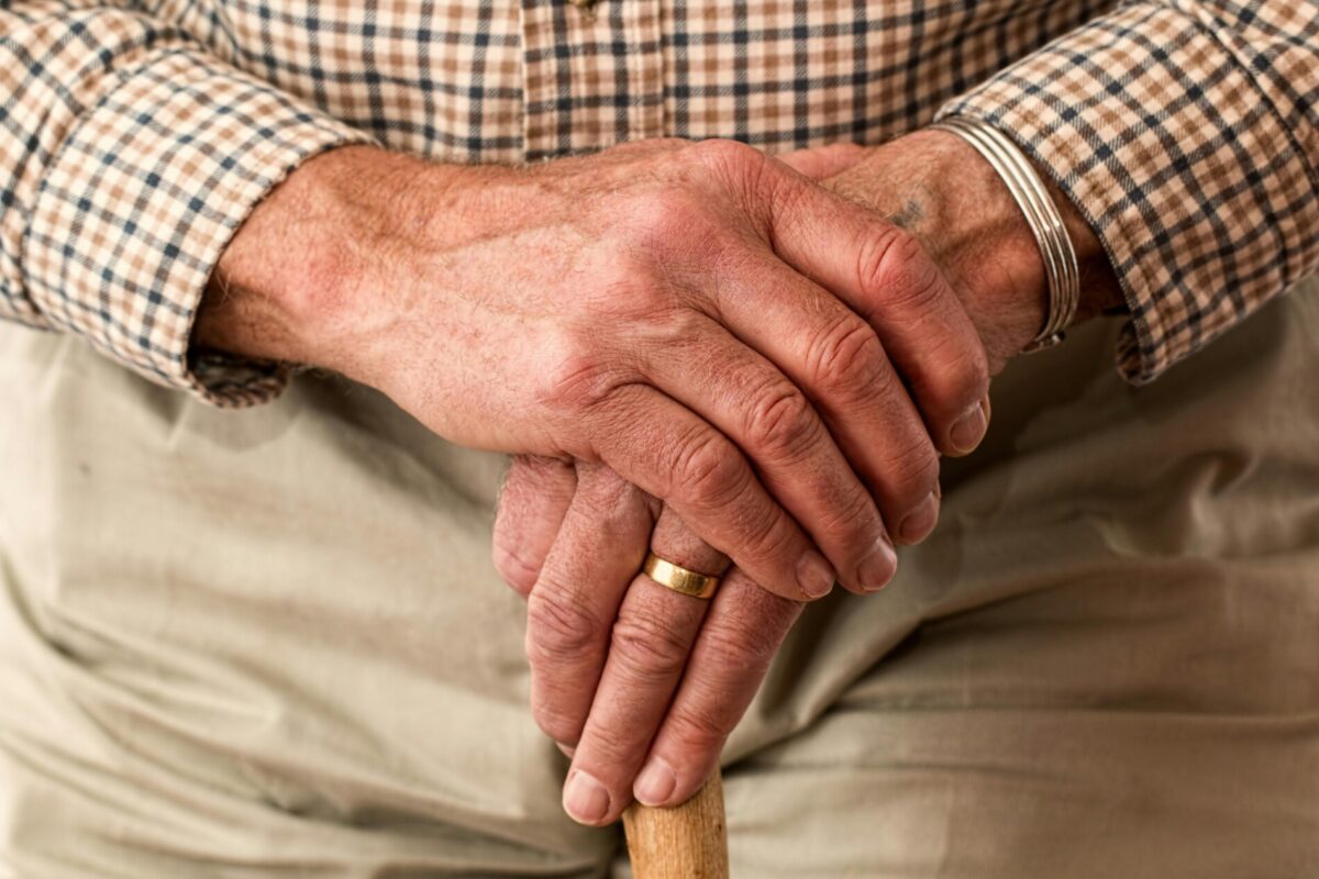 Close-up of elderly person's hands resting on a cane, wearing a plaid shirt and a gold ring.
