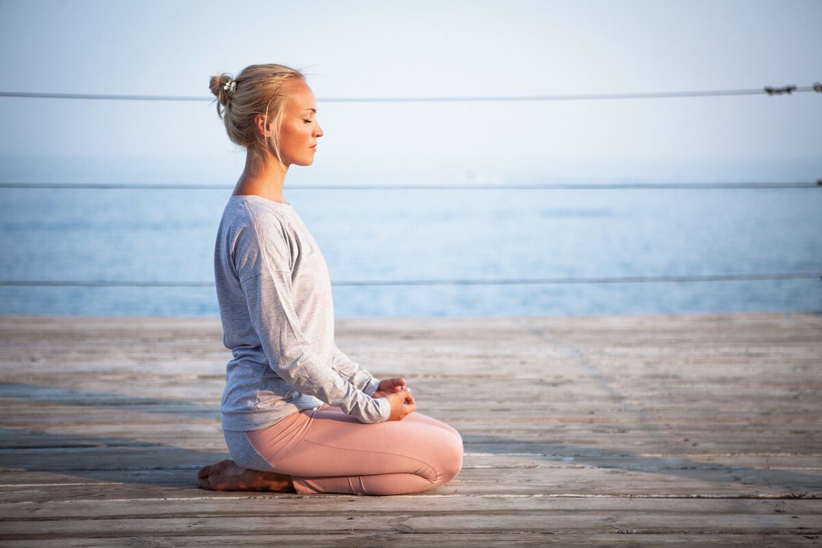 Woman sitting in a kneeling meditation pose on a wooden deck by the ocean, practicing mindfulness with a calm expression.
