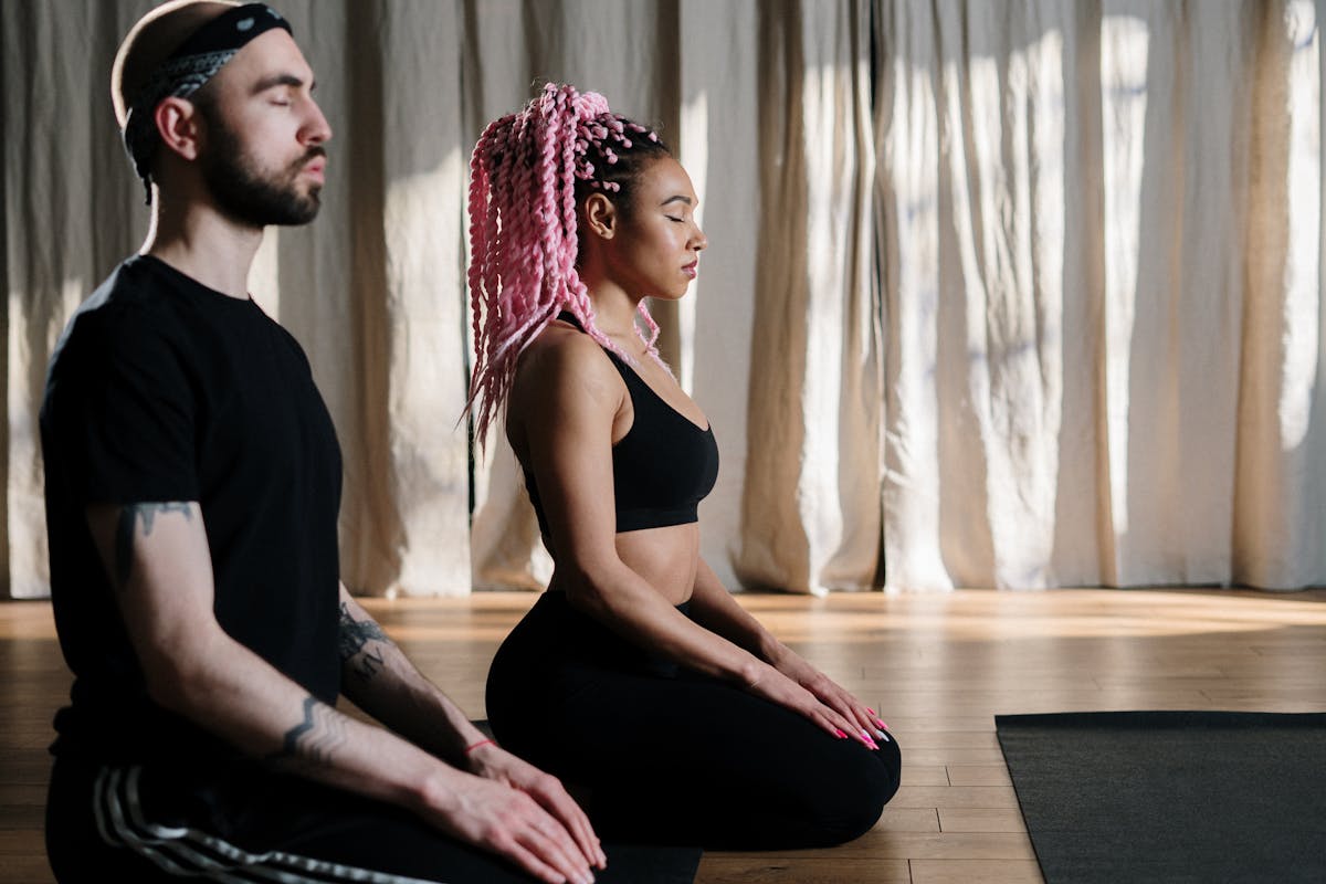 Man and woman sitting in a kneeling meditation pose indoors, with eyes closed and hands resting on their knees, practicing mindfulness.