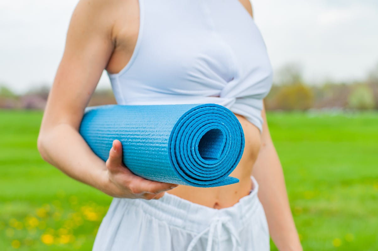 Woman holding a rolled blue yoga mat outdoors, wearing a white top and pants.